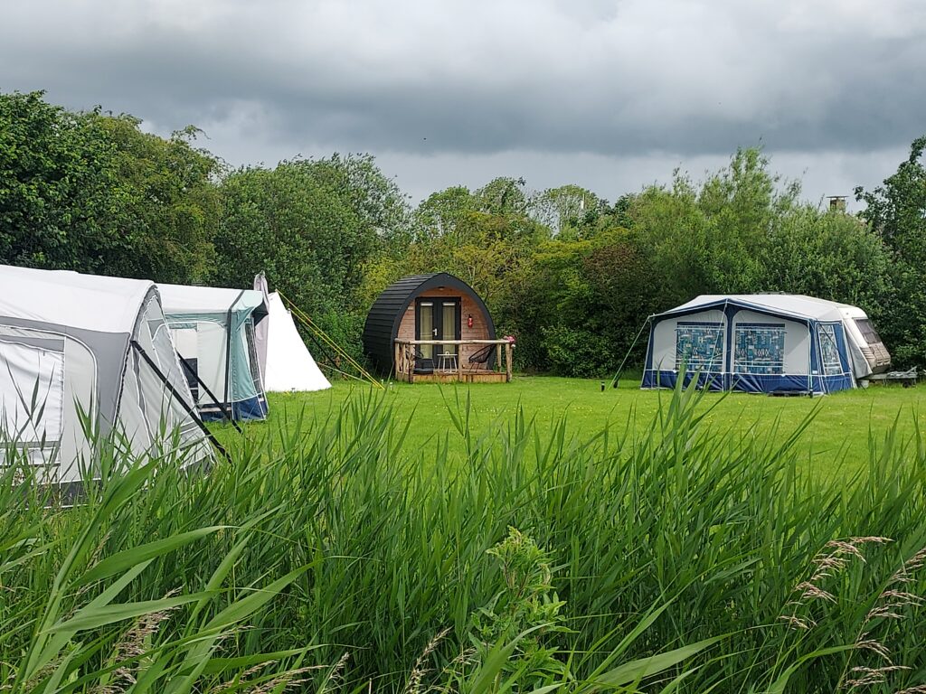 Tweepersoons trekkershut op kampeerveld met caravans en tenten. De kampeerpod heeft een terras met tafeltje en twee loungestoelen. In een mooie groene omgeving.